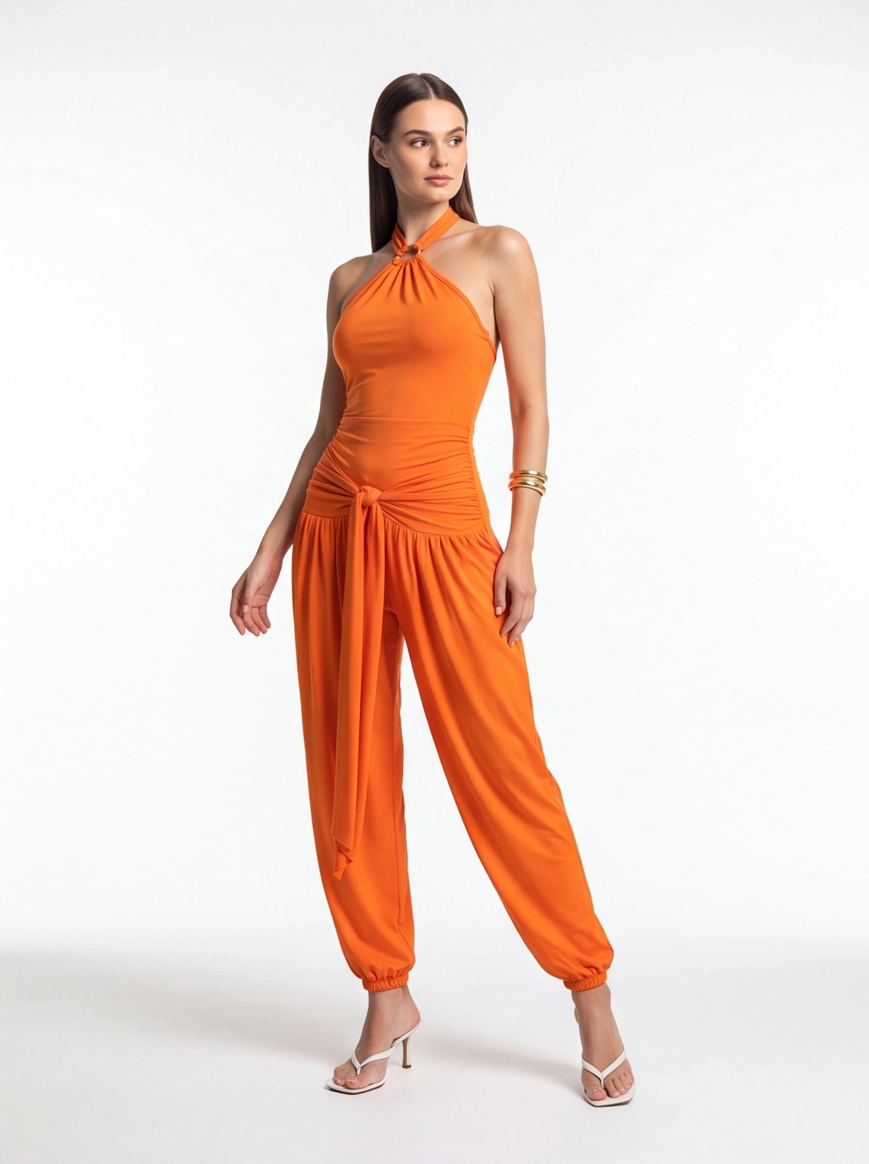 Orange halter jumpsuit with twisted neckline, ruched waist, and waist-tie knot; wide leg with gathered cuffs.