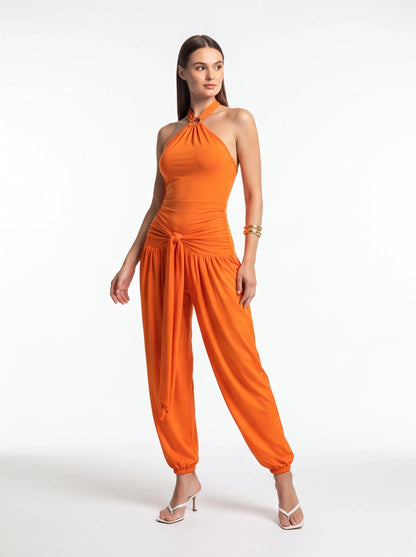 Orange halter jumpsuit with twisted neckline, ruched waist, and waist-tie knot; wide leg with gathered cuffs.