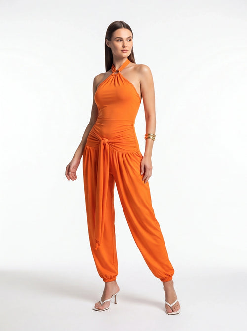 Orange halter jumpsuit with twisted neckline, ruched waist, and waist-tie knot; wide leg with gathered cuffs.
