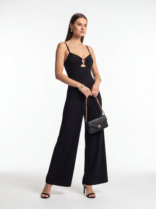 Jamota black jumpsuit with adjustable straps, front ring detail and wide-leg silhouette, paired with a quilted handbag.