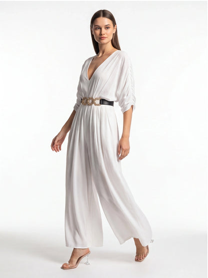 Jamota white jumpsuit with wide legs and deep V-neck, cinched by a black belt with a gold chain.