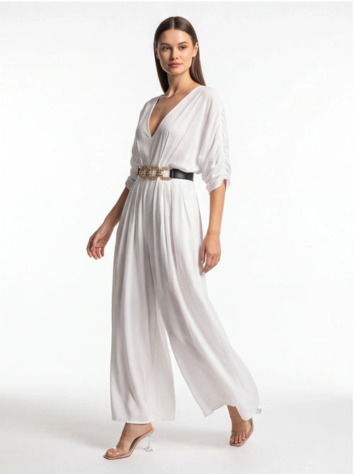 Jamota white jumpsuit with wide legs and deep V-neck, cinched by a black belt with a gold chain.